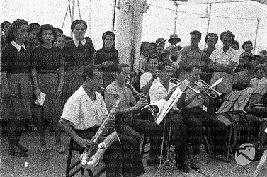 Musicisti con strumenti a fiato tengono un concerto sul ponte di una nave accompagnati da un coro femminile, bambine e bambini in divisa ascoltano in piedi