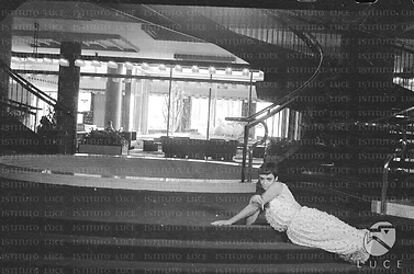 Barbara Steele semidistesa su alcuni gradini all'interno dell'Hotel Hilton. Sullo sfondo, una scalinata circolare; campo medio