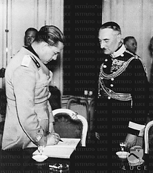 Vienna Galeazzo Ciano (in uniforme di ufficiale della Milizia) scrive su un quaderno sotto lo sguardo di un ufficiale austriaco (?) in alta uniforme