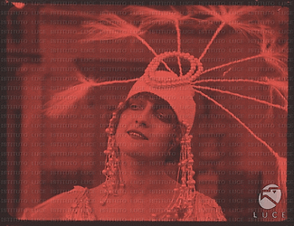 L'attrice Lyda Borelli in una ripresa del film "Carnevalesca" del 1917. L'attrice indossa un copricapo, tipico del film, molto appariscente e con lunghe piume finali. La scena è colorata di rosso.