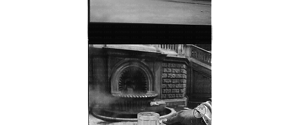 La mano di un uomo tiene un bicchiere d'acqua della  fontana visibile sullo sfondo - primo piano
