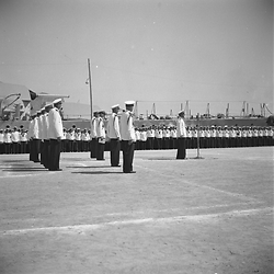 Celebrazione militare in porto: un alto ufficiale parla al microfono, marinai e ufficiali schierati lungo il molo, sullo sfondo si vede la sagoma della nave