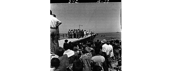 Una piccola folla di bagnanti assiste al passaggio su un molo di Gronchi seguito da un gruppo di autorità; campo lungo