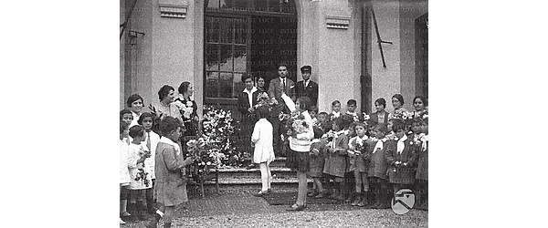 Bambini, bambine e maestri -tutti con dei fiori in mano - sono davanti all'ingresso di una scuola elementare. Una adolescente sta eseguendo il saluto fascista alla volta di una signora. Campo medio