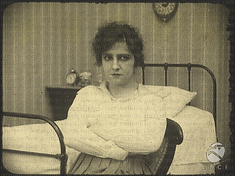 L'attrice Maria Jacobini è seduta su un letto con le braccia conserte e il volto rabbuiato. Indossa una camicia bianca e una gonna con larghe pieghe.