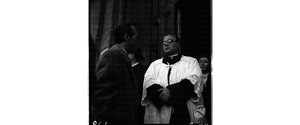 Momenti del funerale di Guido Notari: un religioso attende l'arrivo della bara davanti alla chiesa mentre parla con un uomo - piano americano