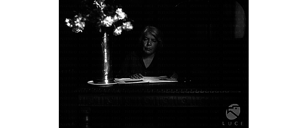 Grazia Deledda seduta alla scrivania in uno studio. Un vaso con fiori sul tavolo. La scrittrice legge un libro.  Medio primo piano