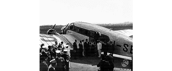 Roma Amelia Earhart colta con un mazzo di fiori vicino alla scaletta dell'aereo; al suo fianco si riconoscono l'ambasciatore Garrett e il marito George P. Putnam