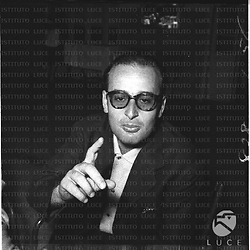 Ritratto di un regista (Luciano Emmer?), seduto al tavolo di un bar, che ha l'indice della mano destra levato verso l'obiettivo della macchina fotografica. Piano medio frontale