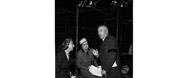 Un uomo anziano, con al braccio una fascia nera, che discute con due donne sotto le strutture in ferro del palco. Piano americano