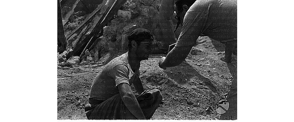 Serge Reggiani accovacciato per terra in mezzo alle macerie, durante la lavorazione del film Tutti a casa. Un uomo gli mostra un oggetto