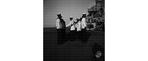 Un alto ufficiale passa in rassegna un gruppo di marinai schierati sul molo di un porto, lato si intravede la nave Bersagliere attraccata