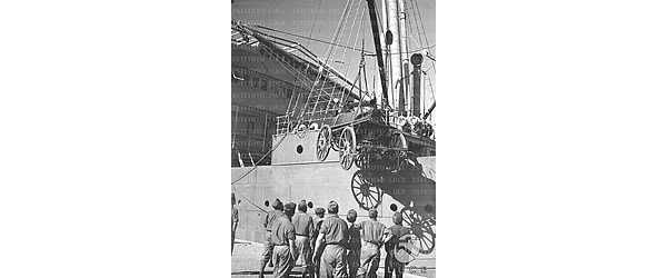 Soldati italiani impegnati in operazioni di scarico di materiali bellici da una nave attendono che una gru cali sul molo una caldaia da campo