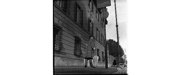 Una modella con un cappotto e un ombrello in mano su un marciapiede di una strada di Roma. Campo lungo