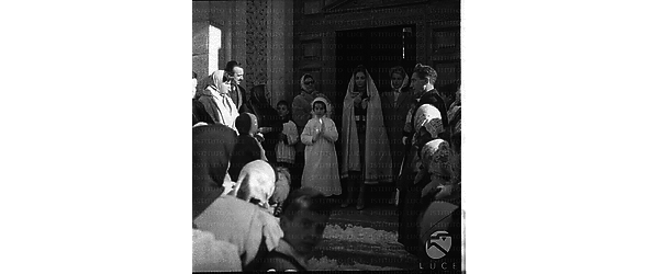Una bambina vestita da Prima Comunione all'uscita da una chiesa, insieme ad altre persone. Tra queste, una donna con un lungo velo - campo medio