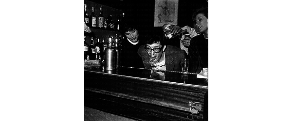 Peppino Di Capri al bancone del bar con le mani dietro la schiena prende un bicchiere con la bocca; accanto a lui Lilly Lembo ed un'altra donna