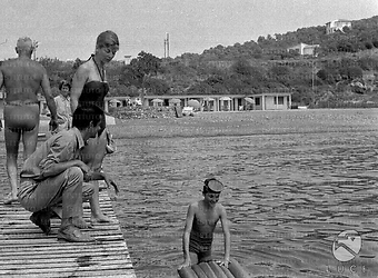 Serge Reggiani su un molo a passerella con la moglie Annie Noel. Il figlio Stephan è in mare, appoggiato a un materassino