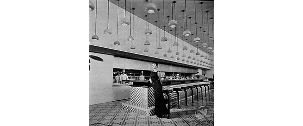 Barbara Steele, in abito da sera, appoggiata al lungo tavolo del bar-self service dell'Hotel Hilton. Visibile l'arredo e i lampadari del salone; campo lungo
