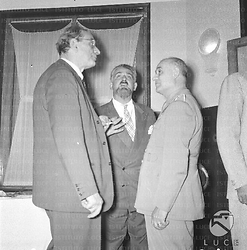 Tre invitati, uno dei quali in divisa, parlano vicino ad una finestra; piano americano