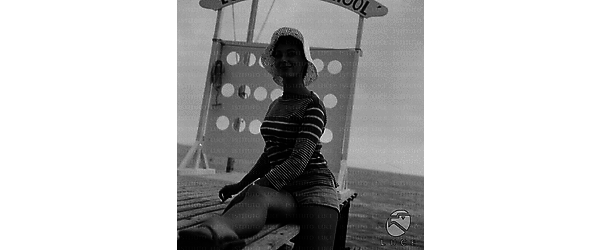 Rita Gam in posa sul molo  con lo sguardo rivolto all'obbittivo,alle sue spalle parte di un'insegna con la scritta: Ecole ski school; totale