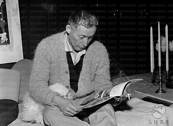 Paul Steffen seduto su una sedia con un barboncino in braccio sfoglia un giornale. Piano americano