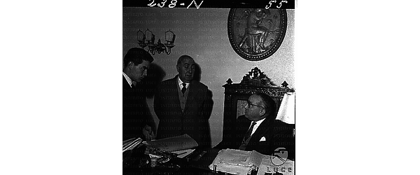Tre uomini, uno dei quali giovane, dietro una scrivania. Campo medio
