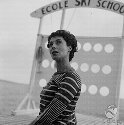 Rita Gam di profilo su un piccolo molo  in legno, alle sue spalle un'insegna con la scritta: Ecole ski school; piano medio