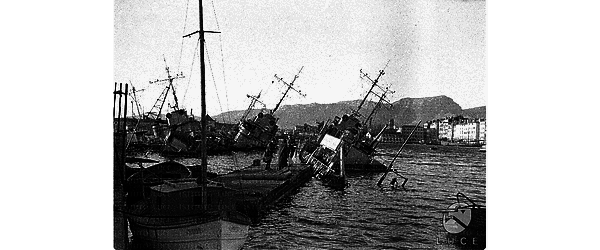 Navi da guerra francesi semi-affondate nelle acque del porto di Tolone, case della città sullo sfondo, persone su un molo galleggiante vicino alle navi semi-affondate