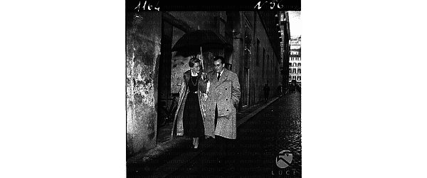 Maria Schell e Luchino Visconti camminano per una via di Roma, coprendosi con l'ombrello - campo medio
