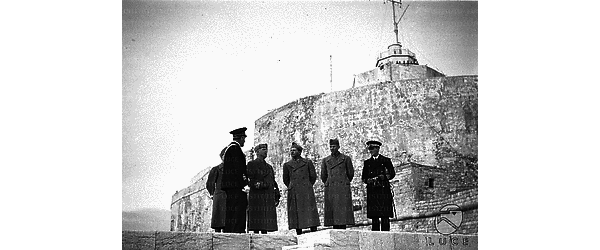 Il re e alcune autorità militari conversano, in occasione della visita alla base militare marittima di Trapani, sullo sfondo del castello de La Colombaia - campo medio - Scorcio dal basso verso l'alto