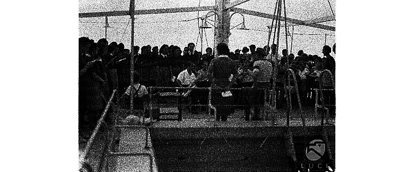 Musicisti con strumenti a fiato tengono un concerto sul ponte di una nave accompagnati da un coro femminile, bambine e bambini in divisa ascoltano in piedi