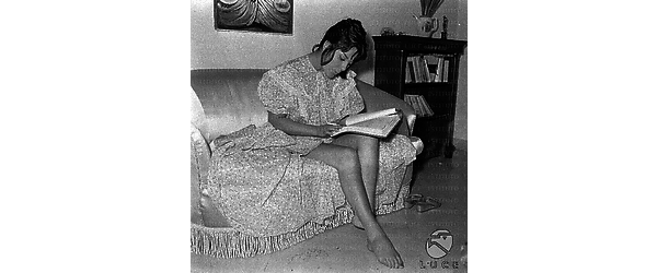 Daniela Rocca seduta su un divano di casa con un libro sulle gambe. Totale