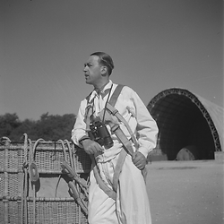 Un militare con la tuta bianca e il binocolo guarda lontano, sullo sfondo si vede un capannone dell'Aeronautica
