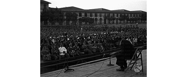 Un attore seduto su una sedia sul palco legge qualcosa, ufficiali tedeschi in prima fila, il pubblico riempie il cortile