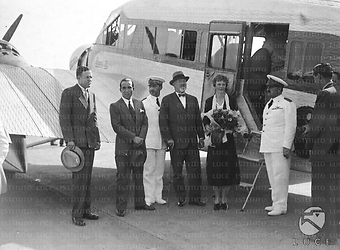 Roma Amelia Earhart colta con un mazzo di fiori vicino alla scaletta dell'aereo; al suo fianco si riconoscono l'ambasciatore Garrett e il marito George P. Putnam
