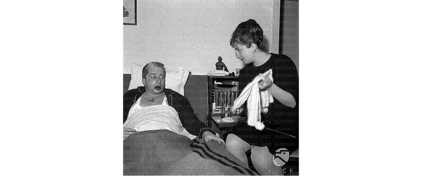 Carlo Croccolo ferito nel suo letto con accanto la moglie che gli mostra un lavoro a maglia che sta facendo