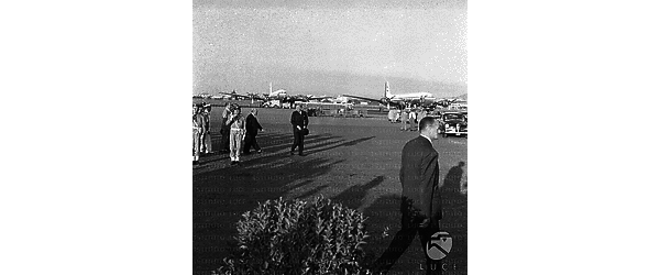 Eden e Piccioni colti sul piazzale dell'aeroporto con truppe schierate. In primo piano un uomo che cammina. Campo lungo