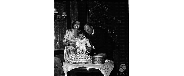 Coppia di genitori - Ing. Brugnoli e moglie (?) - festeggia in casa il primo anno di età della propria bambina: il padre aiuta la piccola a tagliare la torta posta su un tavolino. Totale