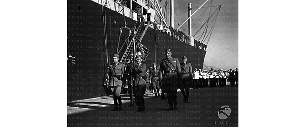 Un gruppo di alti ufficiali italiani sfila lungo un molo di fronte a una nave carica di soldati schierati mentre una banda militare della Marina suona