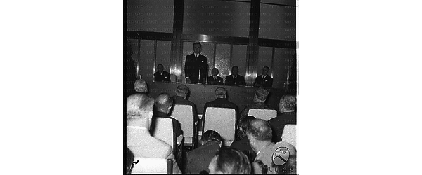 Pella pronuncia un discorso dal palco durante l'inaugurazione del Palazzo della Farnesina. Con lui sul palco Folchi, Segni, Togni e, probabilmente, Rossi. Di spalle l'uditorio - campo medio
