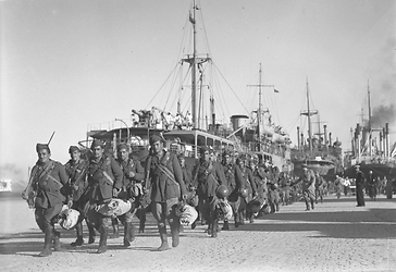 Una colonna di soldati italiani in marcia lungo un molo nel porto di Durazzo, navi sullo sfondo, un marinaio assiste alla scena