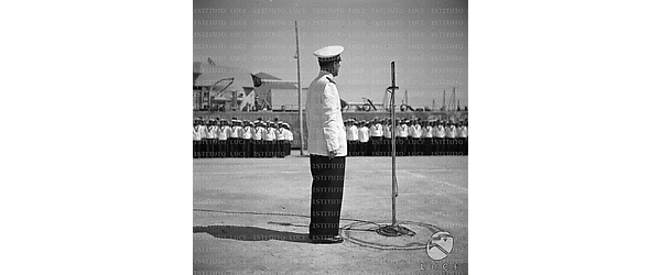 Un alto ufficiale tiene discorso nel molo di un porto di fronte a dei reparti della Marina schierati, sullo sfondo si vede una grande nave da guerra