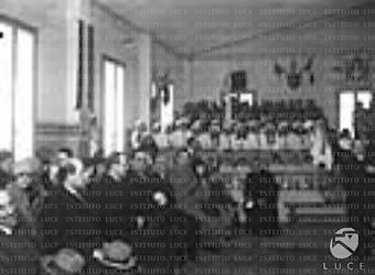 Roma Bottai ed altre autorità seduti in una grande stanza pavesata di bandiere. Adolescenti in uniforme collegiali su un piano rialzato.