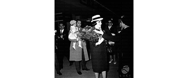 Gina Lollobrigida all'aeroporto riceve un fascio di rose da una donna; dietro di lei il marito con il figlio in braccio - totale