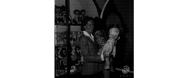 Madeleine Robinson, in un negozio di giocattoli, guarda una bambola sorridendo; piano americano