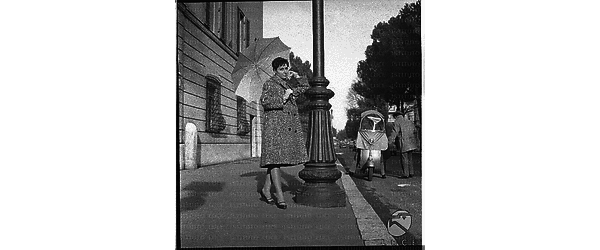 Una modella con un cappotto e un ombrello in mano su un marciapiede di una strada di Roma. Campo medio