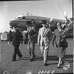 Lord e lady Jowitt, accompagnati da un uomo e da un assistente di volo, si dirigono verso l'uscita dell'aeroporto. Campo medio