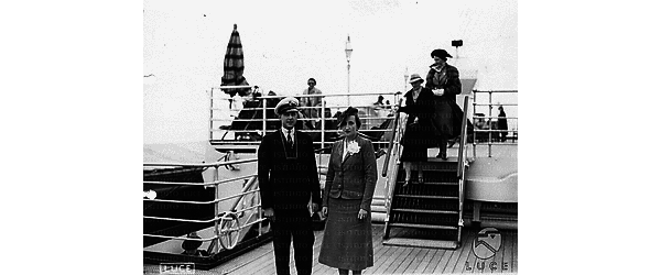 Golfo di Napoli Ritratto sul ponte della nave: una donna con cappellino e fiore bianco all'occhiello della giacca in compagnia di un uomo in divisa; sullo sfondo due eleganti signore scendono le scalette
