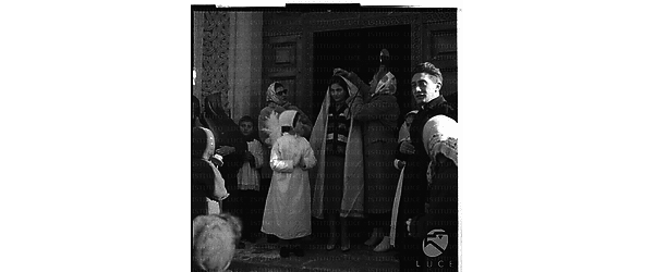 Una bambina vestita da Prima Comunione all'uscita da una chiesa, insieme ad altre persone. Una donna ne copre un'altra con un lungo velo - totale