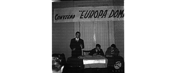 Un uomo in piedi dal tavolo degli oratori tiene un discorso. Alla sua sinistra è seduto il professore Pagliari. Campo medio